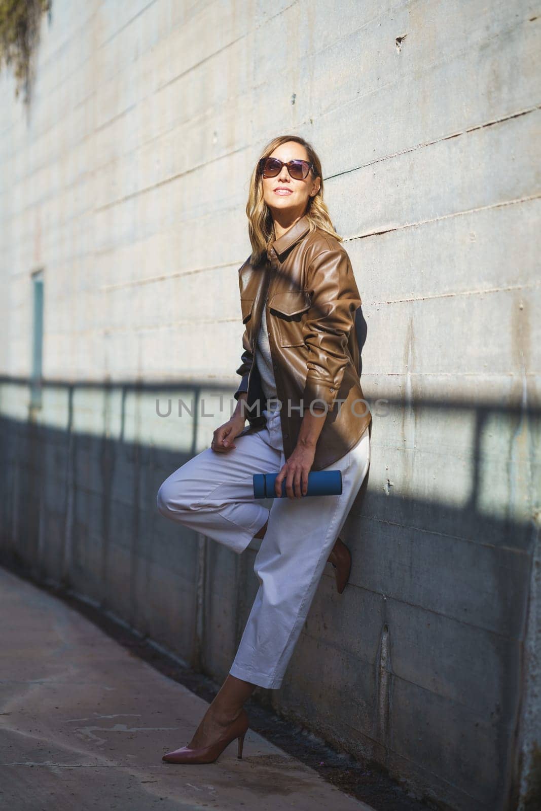 Full body glad woman in stylish clothes with thermos leaning on sunlit wall and looking at camera with smile on weekend day