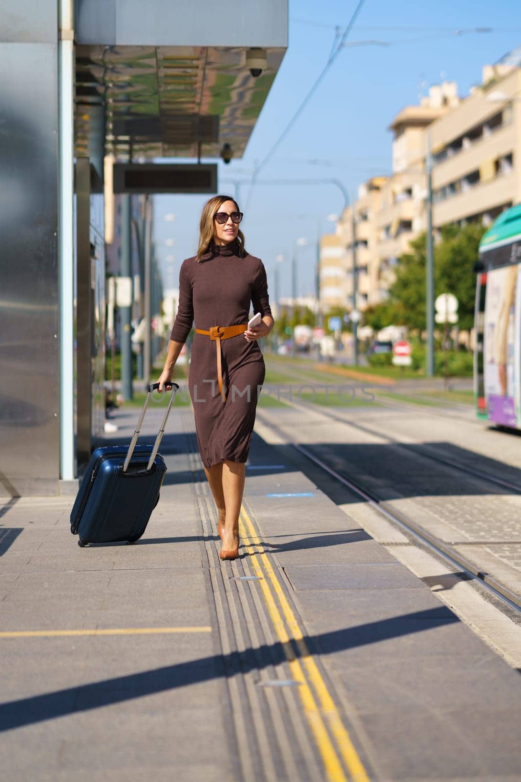Full body stylish female entrepreneur with suitcase and cellphone walking on platform near tramway during business trip in city in daytime