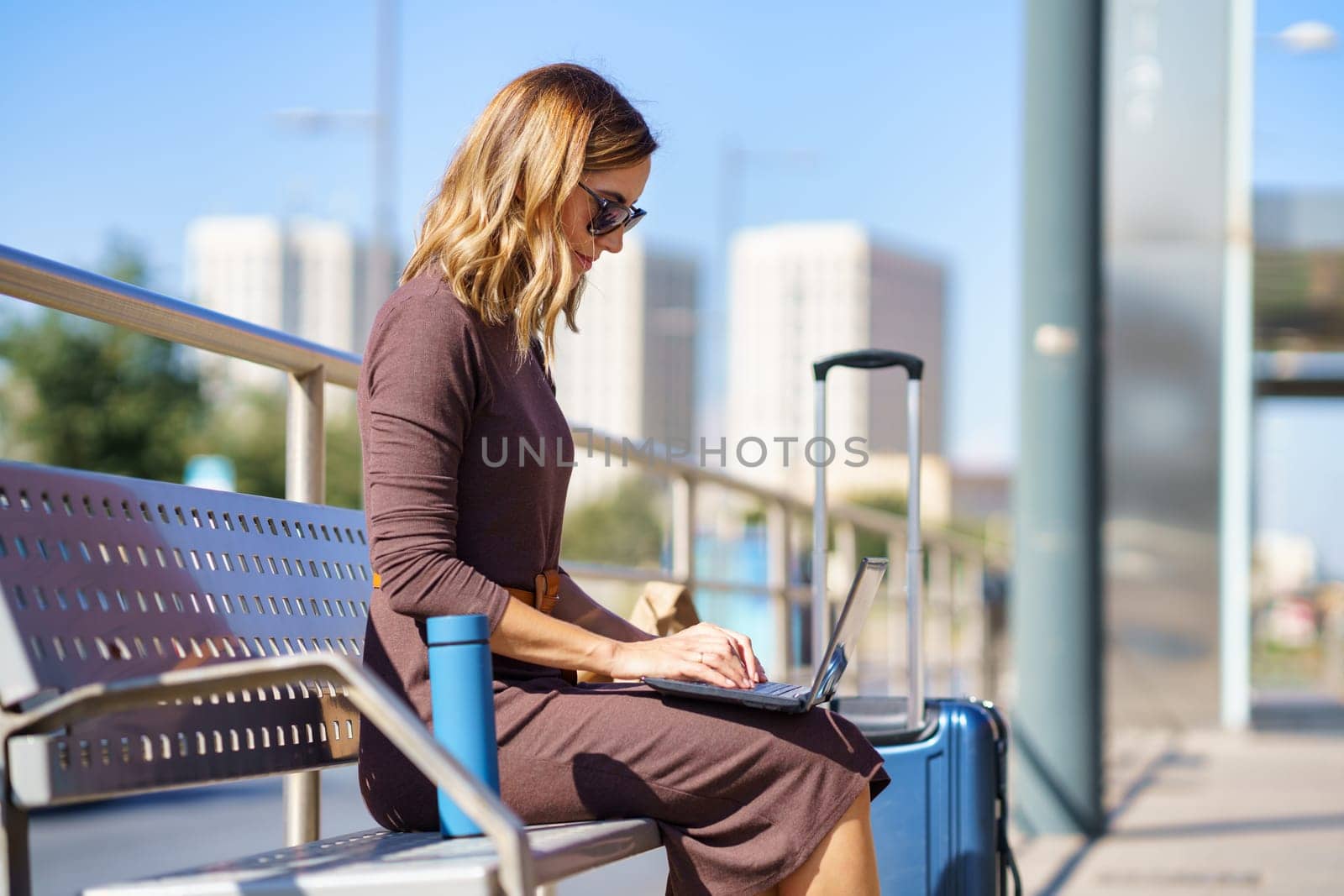 Female manager in stylish dress and sunglasses sitting on bench near suitcase and thermos and browsing netbook on station during business trip in city