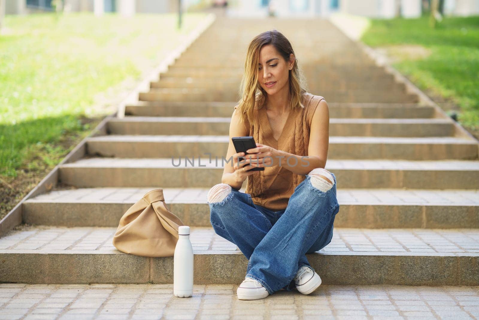 Full body adult female in stylish clothes sitting on stairs near bag and bottle and browsing social media on cellphone on street