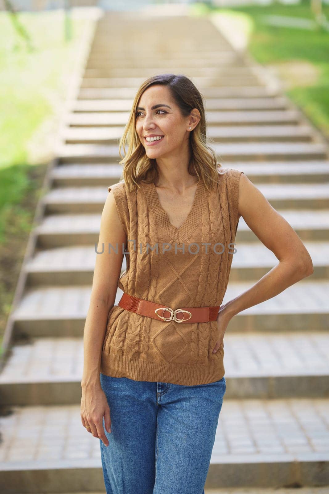 Positive adult female in stylish knitted top holding hand on waist and looking away with smile on blurred background of steps