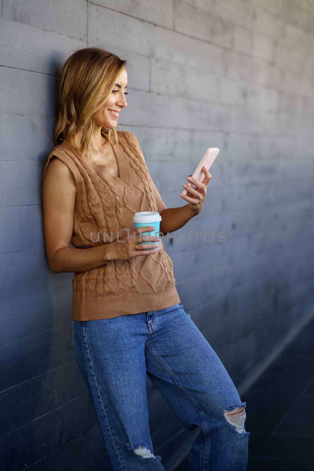 Cheerful woman with smartphone and coffee leaning on wall by javiindy