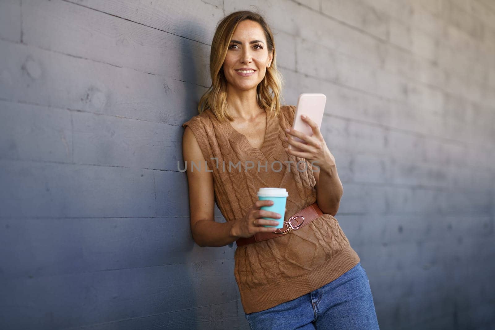Woman in stylish knitted top, with cup of coffee to go looking away with smile and using smartphone while leaning on gray wall on city street