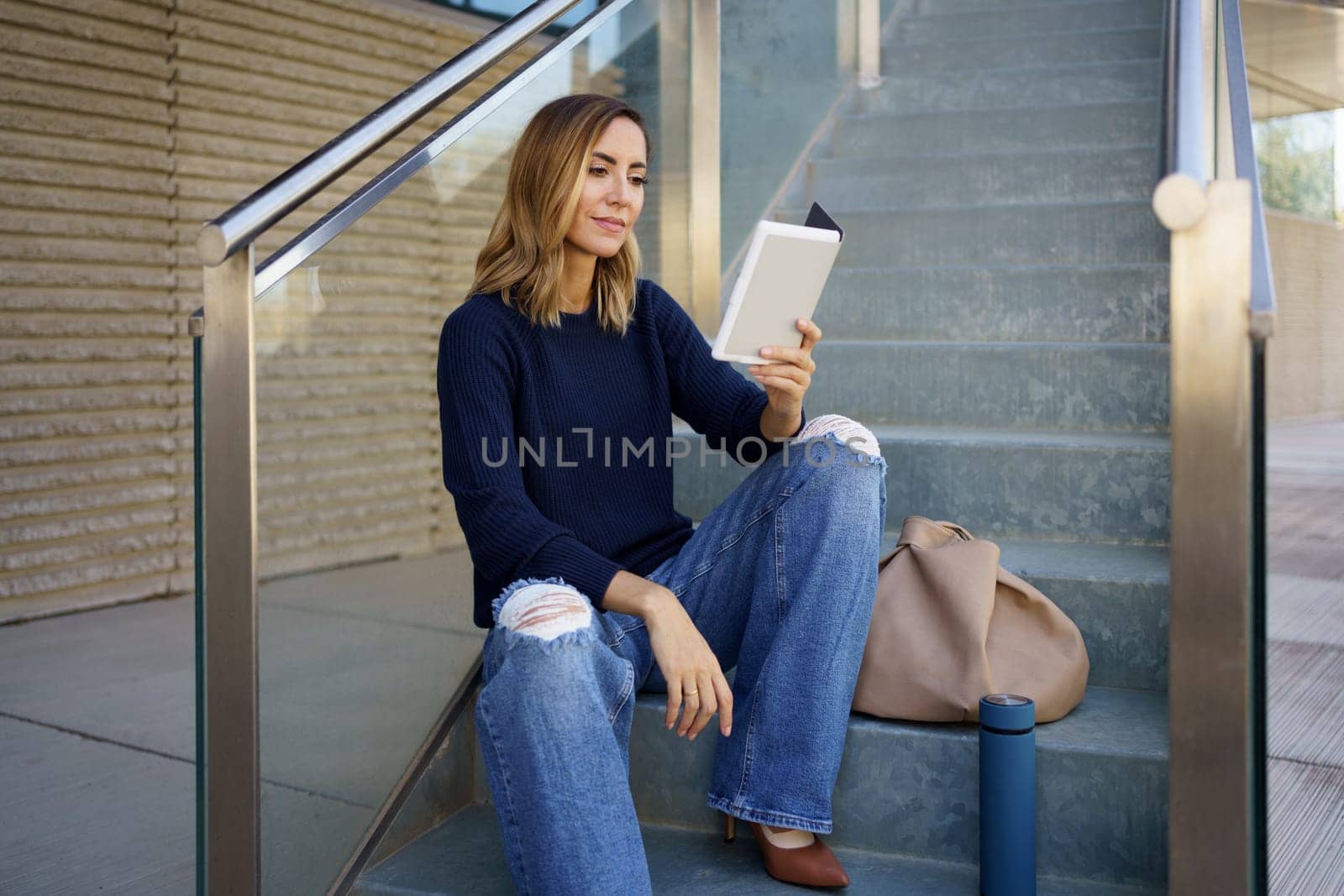 Female in stylish clothes sitting on stairs near bag and reading e book in daytime on city street