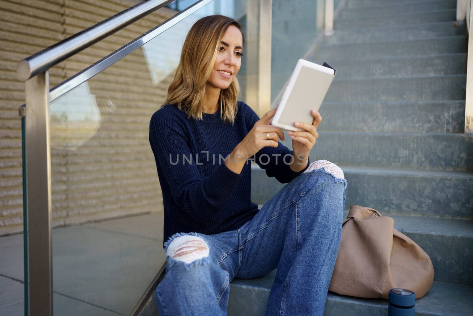 Positive female in stylish clothes smiling and swiping pages of e book while sitting on stairs and reading in daytime on city street