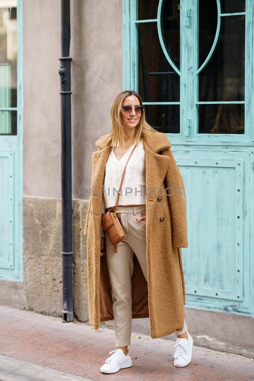 Full body of young female in stylish brown coat sneakers and sunglasses standing on street near building