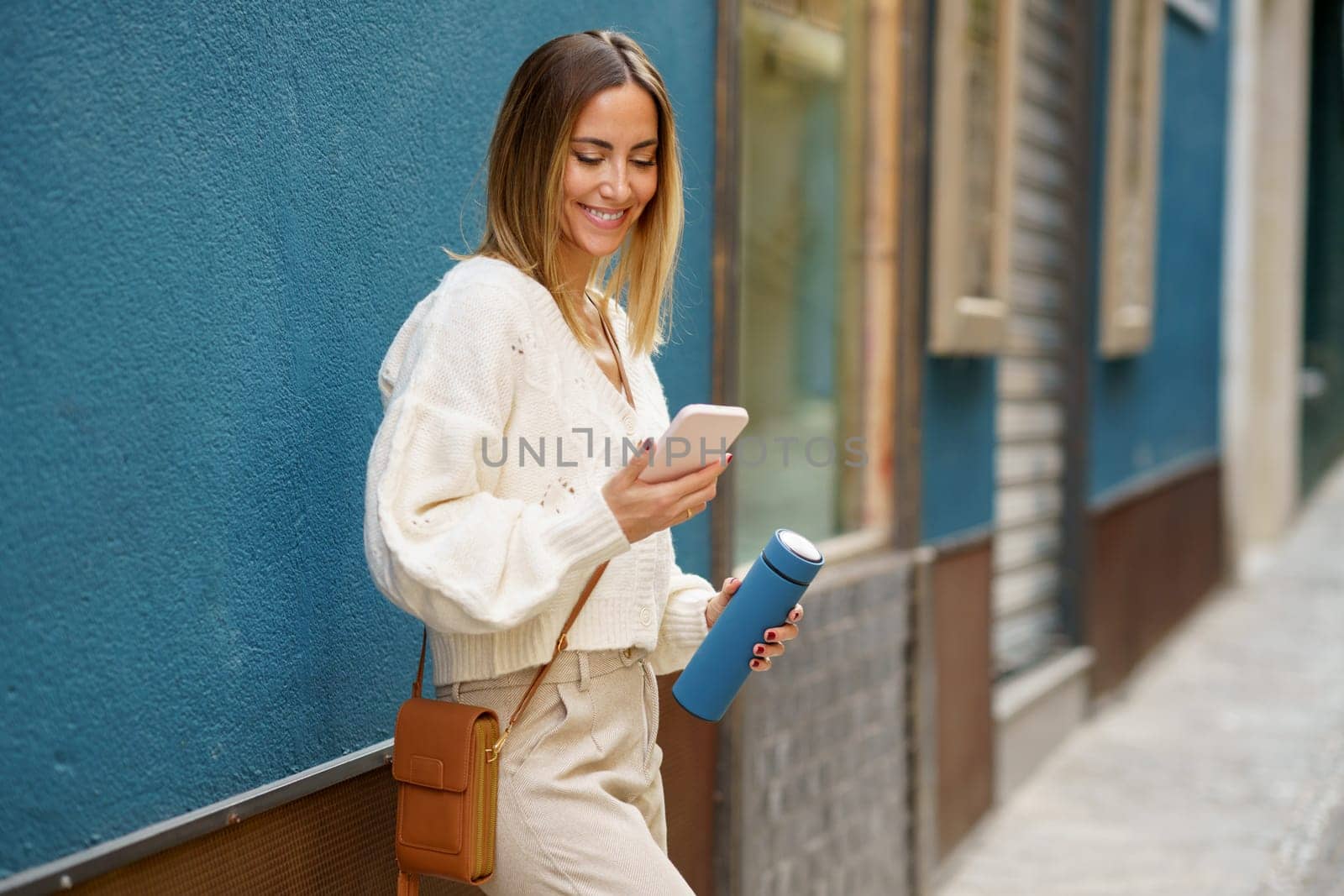 Cheerful young female in smart casual wear standing near blue wall while browsing mobile phone and messaging