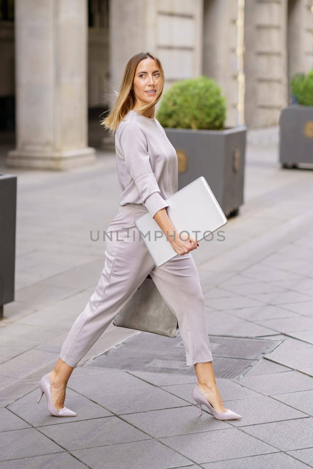 Side view of elegant young feminine businesswoman with long blond hair in stylish outfit, and high heels walking on paved street with laptop in hand and looking away