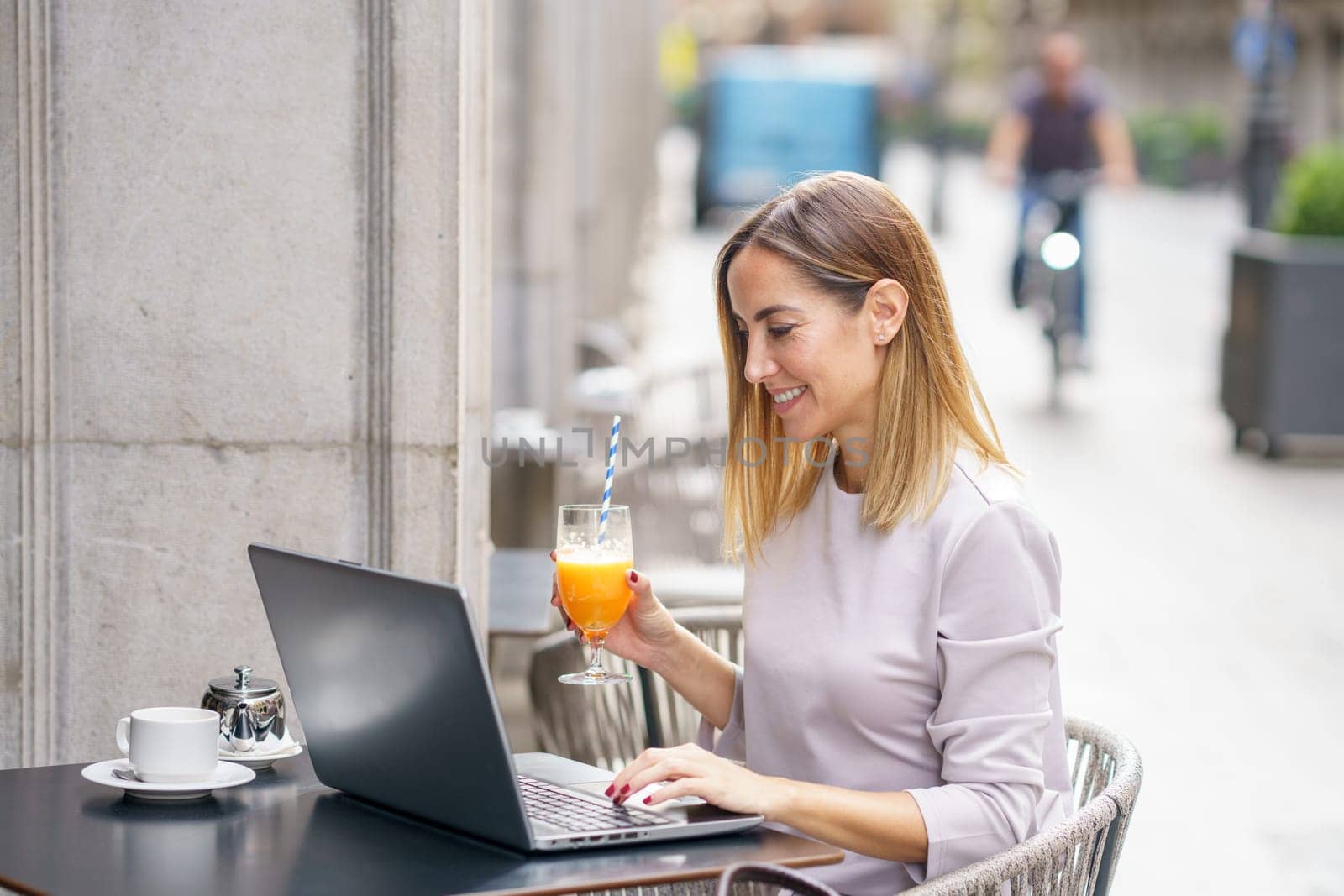 Side view of positive adult self employed lady with blond hair in casual clothes, drinking glass of fresh juice and smiling while working remotely on laptop in street cafe