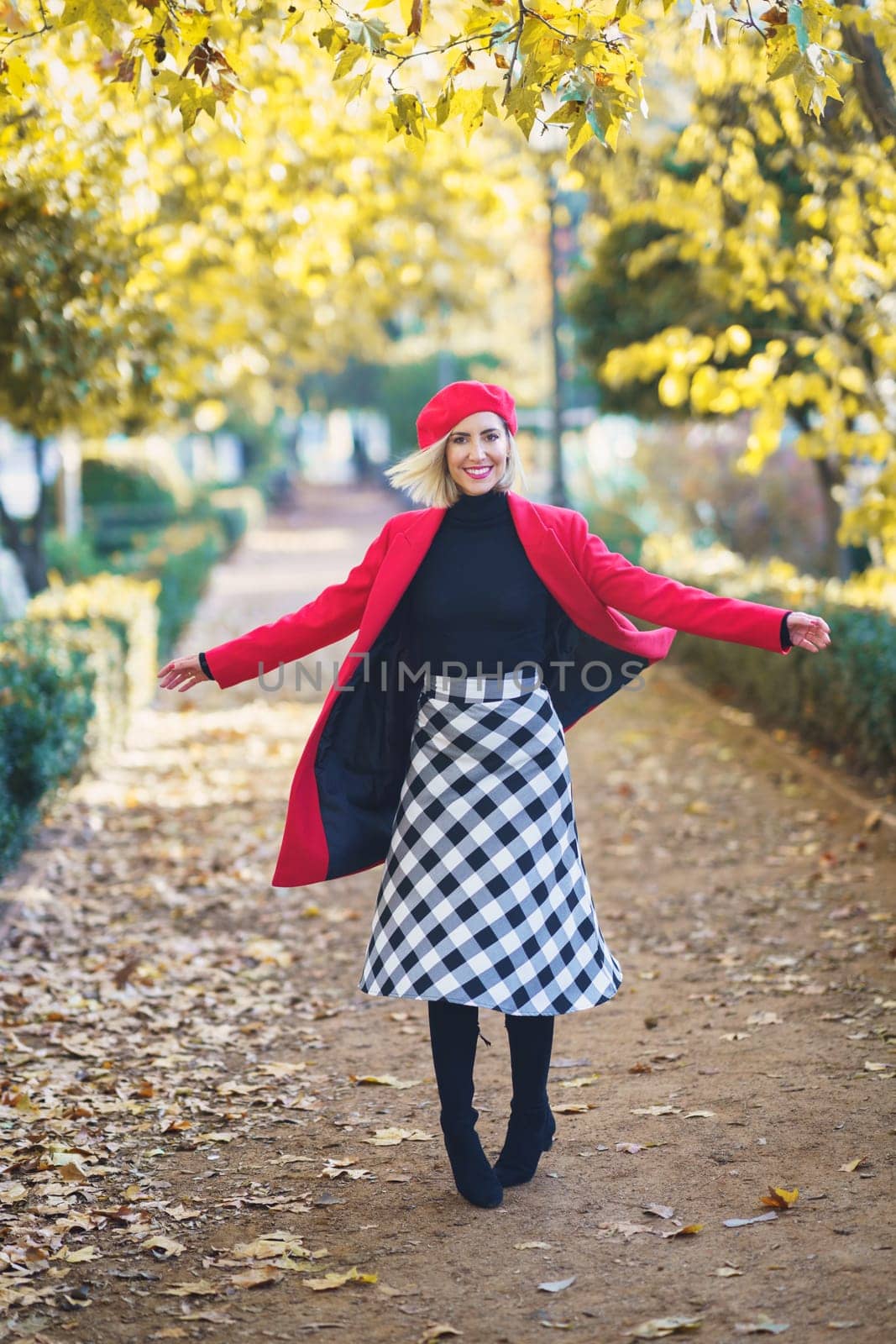 Full body of cheerful female in red coat and beret looking at camera while spinning around on walkway in autumn park