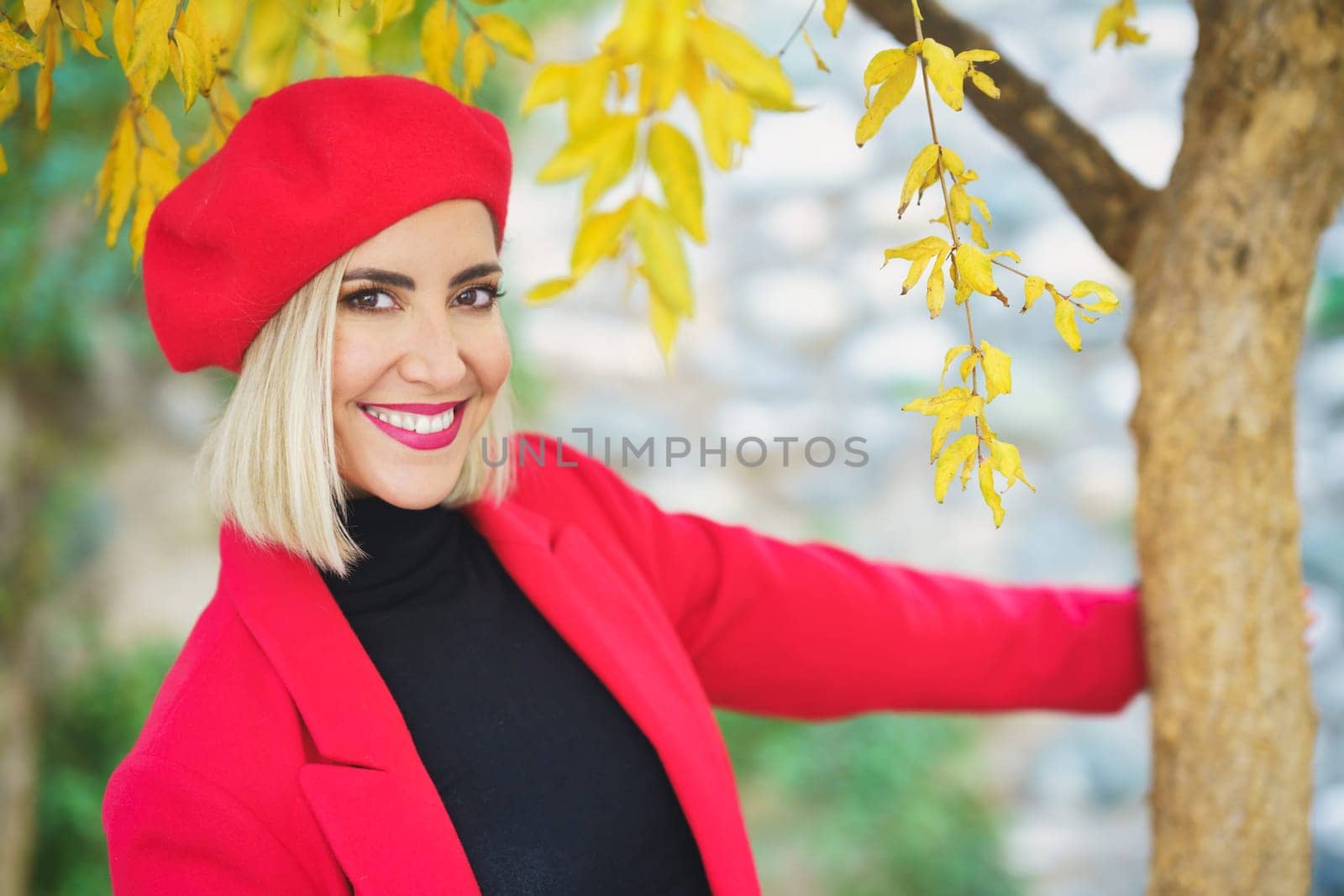 Self assured young content lady, with blond hair and makeup in stylish outfit and beret smiling brightly and looking at camera while touching tree trunk in autumn park