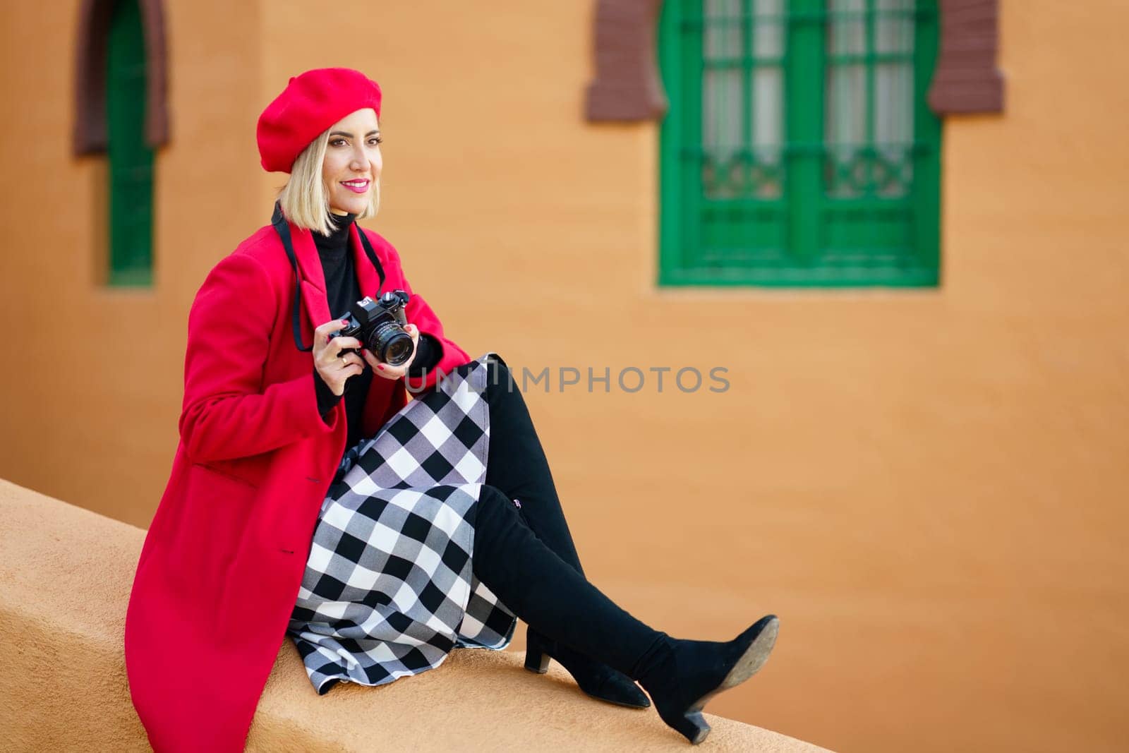 Middle-aged woman wearing red winter clothes, taking pictures with an SLR camera sitting on a city wall. Female wearing coat, skirt and beret outdoors.