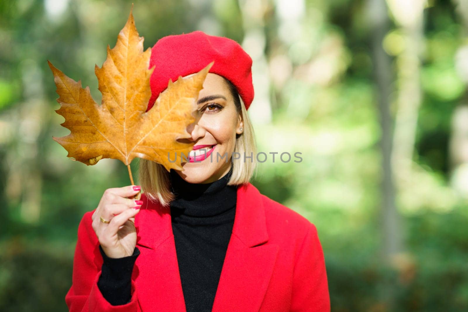 Delighted female in red beret and coat hiding half of face behind dry autumn leaf in sunny park and looking at camera
