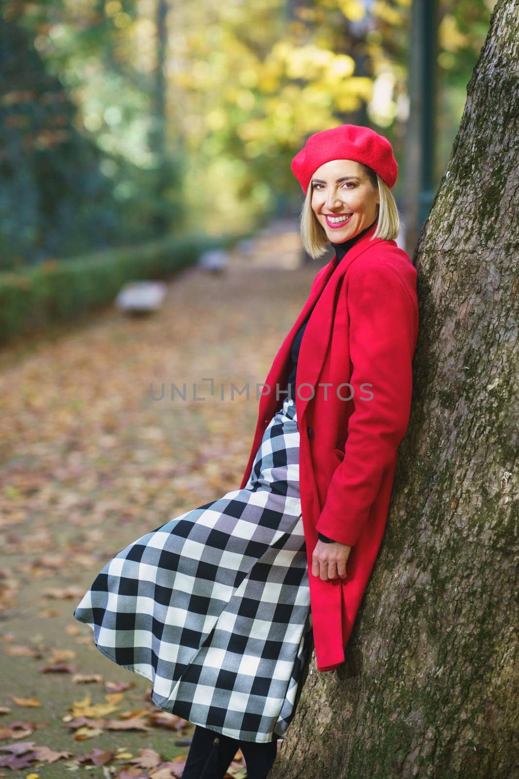Side view of smiling female in elegant outfit leaning on tree on walkway in autumn park and looking at camera