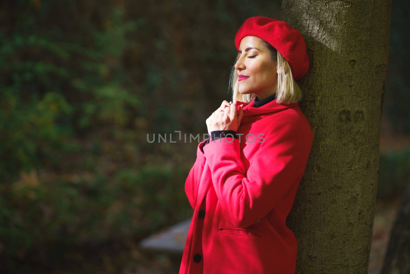 Side view of serene female with closed eyes adjusting collar of red coat while standing in sunlight near tree in park