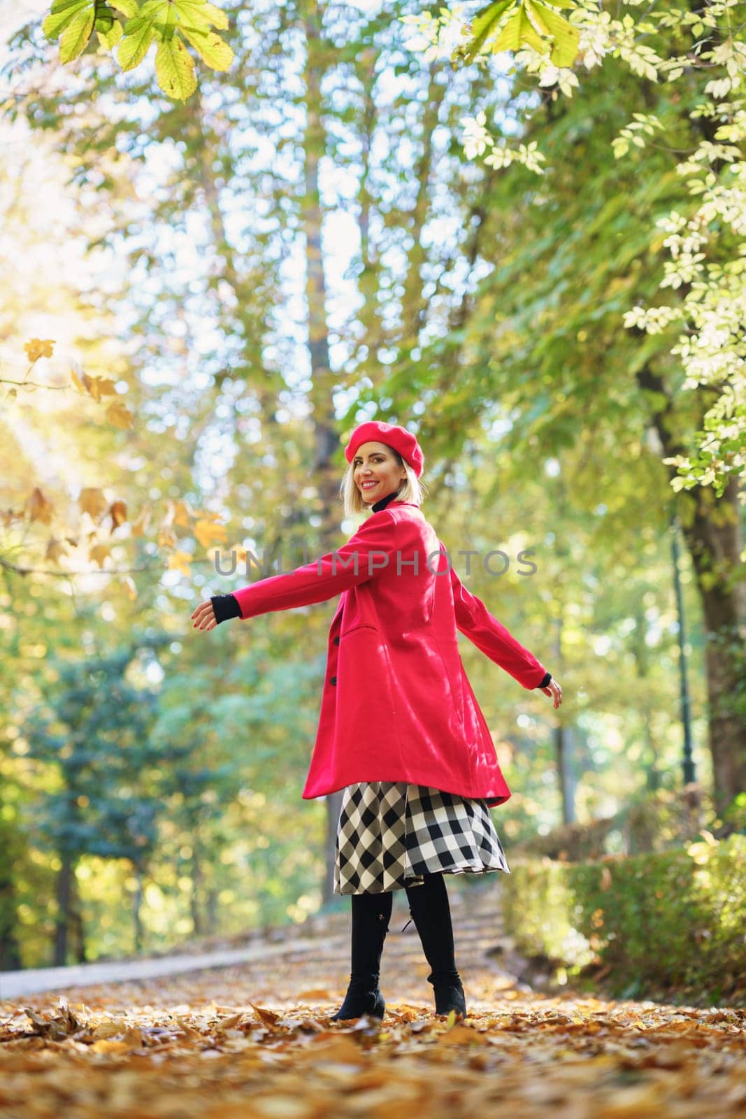 Full body side view of positive female in red clothes spinning around while standing on alley with dried leaves in autumn park