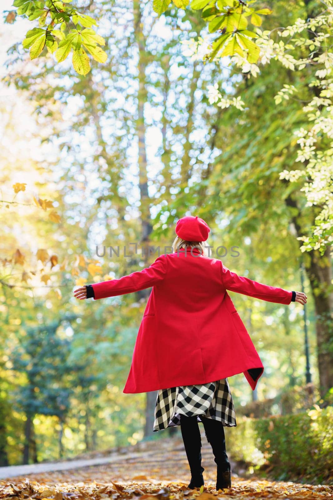 Full body back view of anonymous female in red outfit standing with spread arms on path with fallen leaves in autumn park