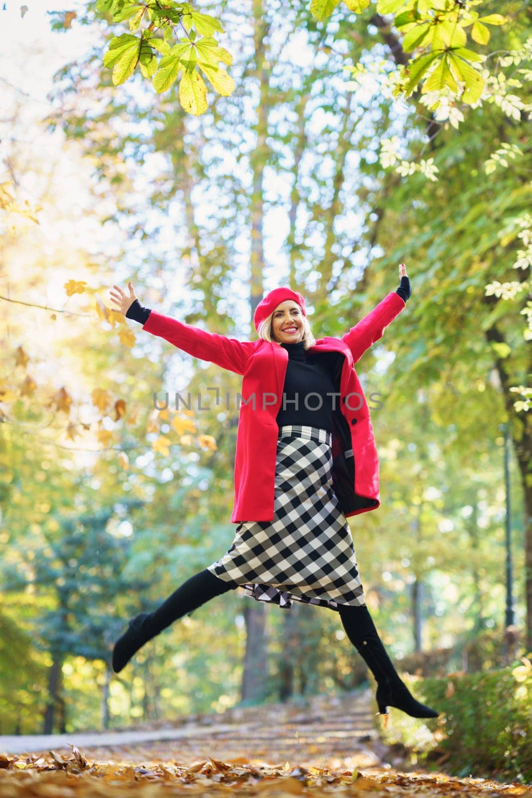 Full body of positive female in bright coat and checkered skirt looking at camera with spread arms while jumping in park on autumn day