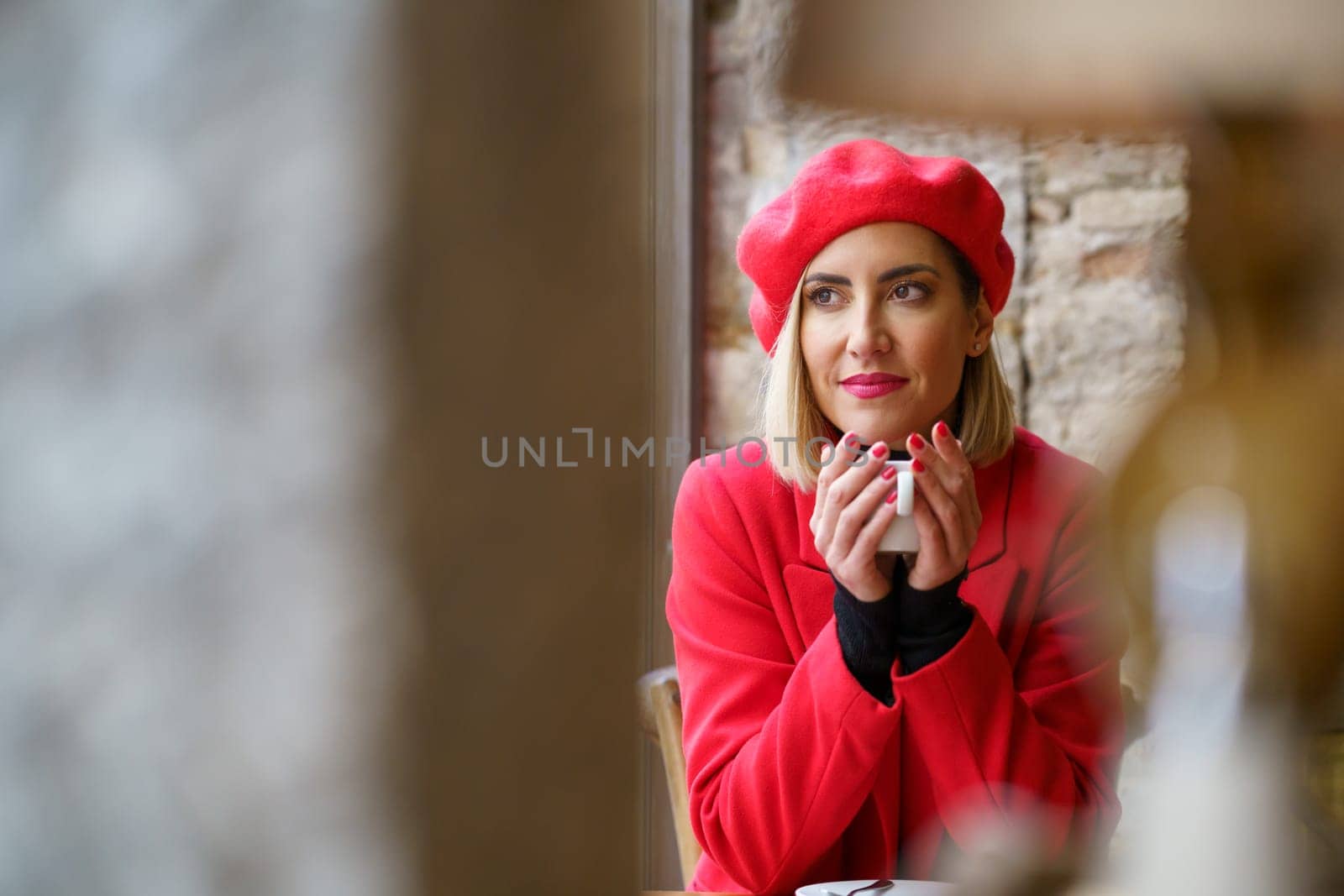 Through blurred column and lamp of charming female in red clothes looking away while sitting with cup of hot drink in modern cafeteria