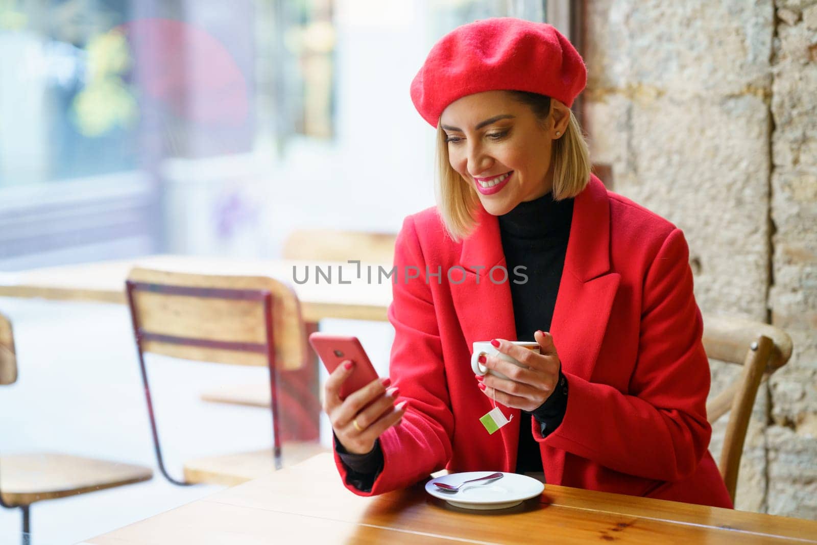 Cheerful female in stylish red outfit scrolling social media on cellphone while sitting at table with cup of tea in cafeteria