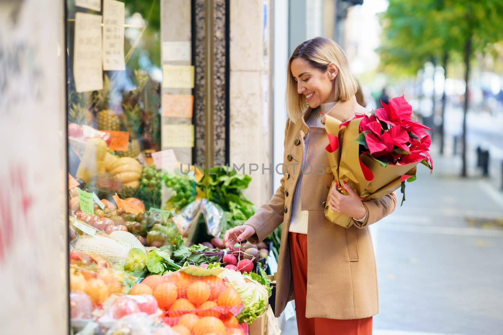 Side view of smiling female with bouquet of flowers buying fresh vegetables while standing at stall on street of city