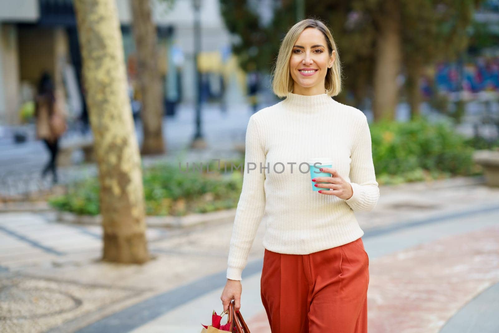 Positive female wearing turtleneck and pants walking with bag and holding cup of takeaway coffee drink in park