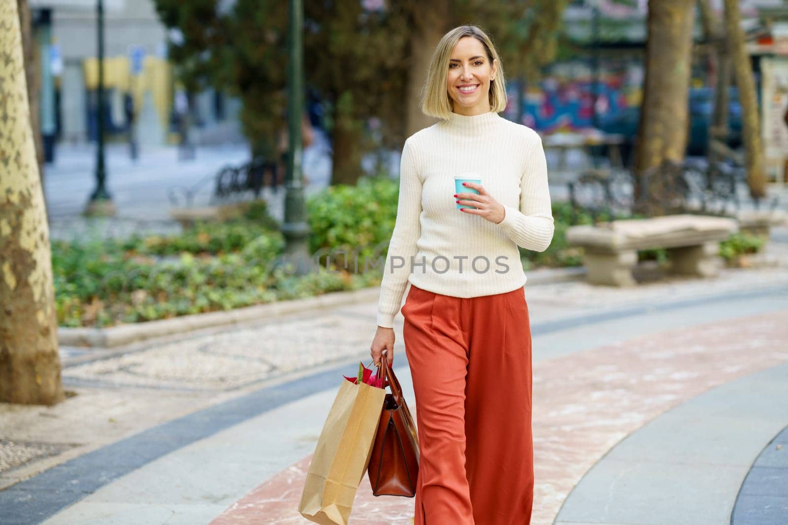 Positive female in casual clothes carrying shopping bag and handbag and having cup of takeaway beverage in park