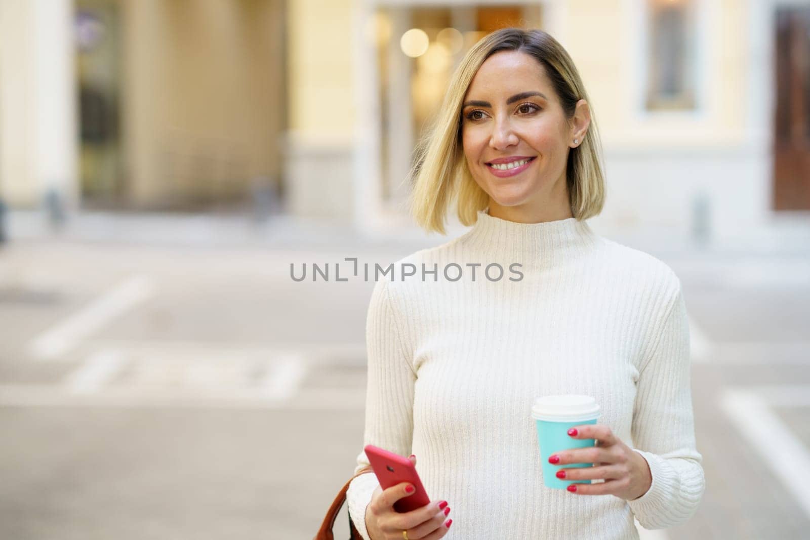 Smiling woman with takeaway coffee and smartphone on street by javiindy