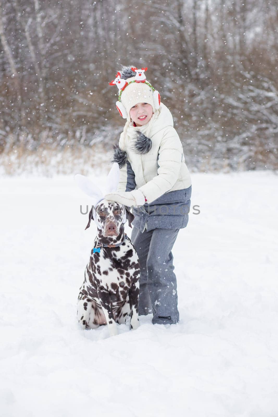 Girl wearing winter warm coat, hat and Christmas headband has fun with dalmatian dog outdoor. new year card, girl walking with dog in winter forest. funny childhood. by YuliaYaspe1979