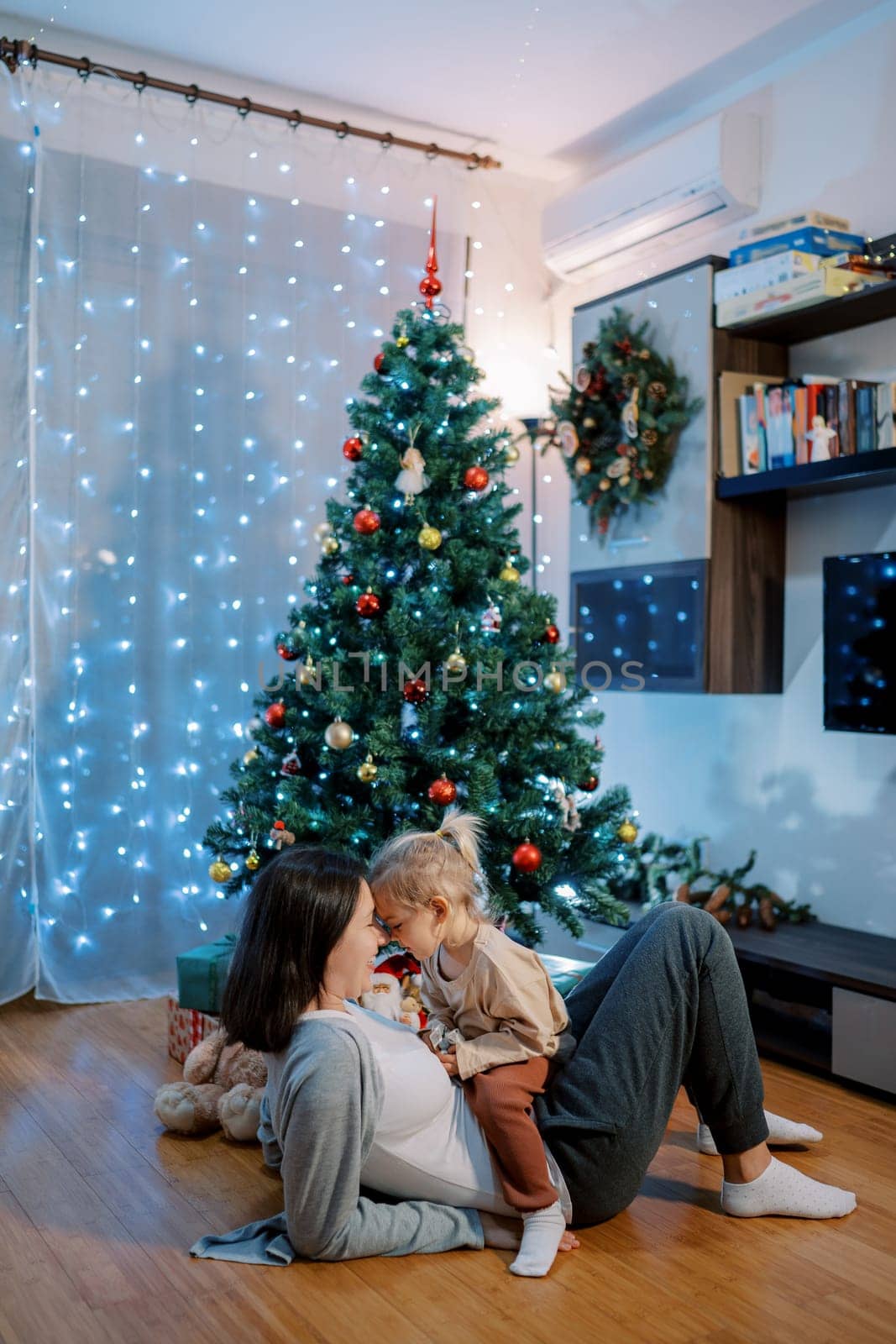 Little girl sits on her mother stomach, touching foreheads with her on the floor by the Christmas tree. High quality photo