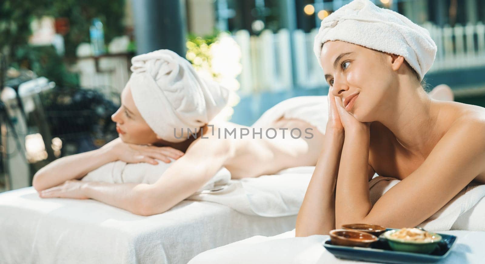 A couple of beautiful young woman relaxes on a spa bed surrounded by nature at outdoor. ready for a body massage. Attractive females in white towels lie on bed peacefully. Close up. Tranquility