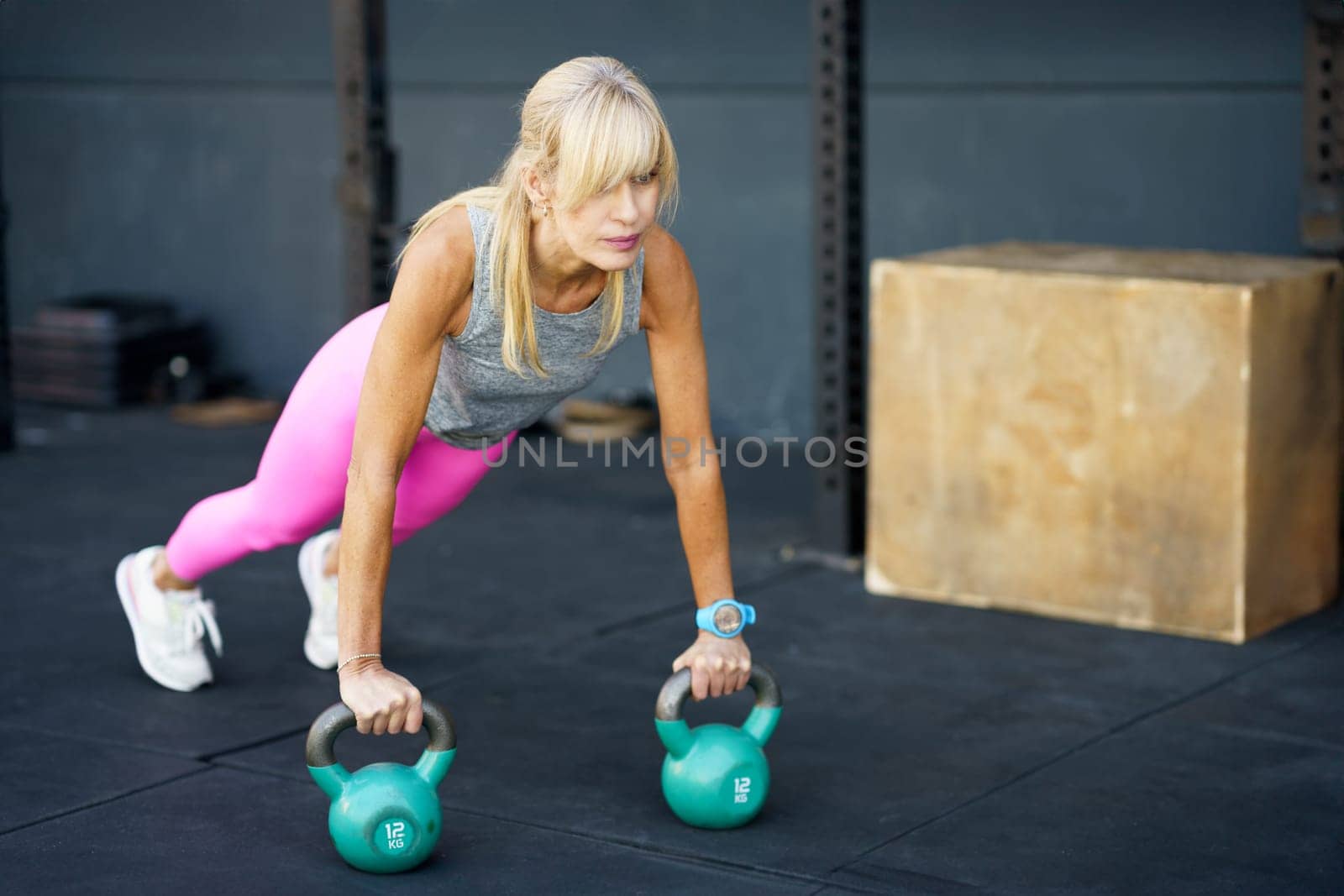 Fit strong blonde hair in activewear doing push ups exercise with heavy kettlebells during functional fitness workout at modern gym against blurred background