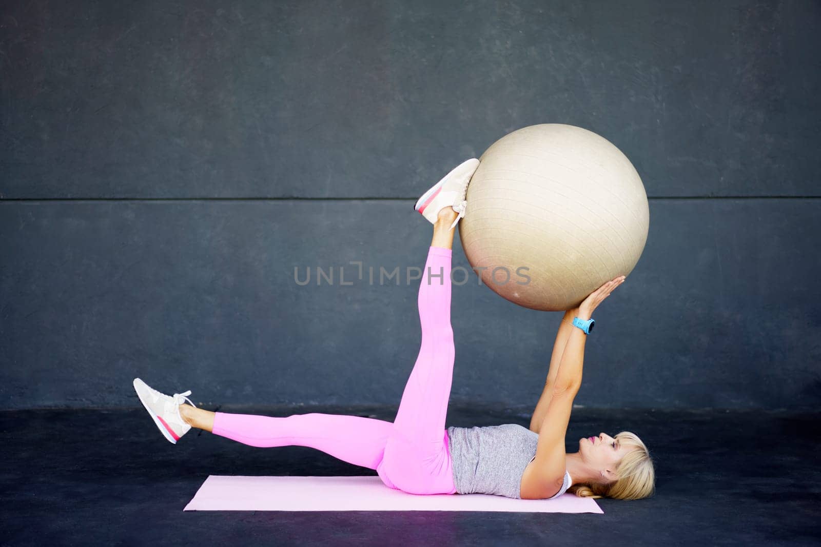 Side view of flexible fit female stretching leg, and doing exercise with fitness ball while lying on mat in modern gym against gray wall during workout