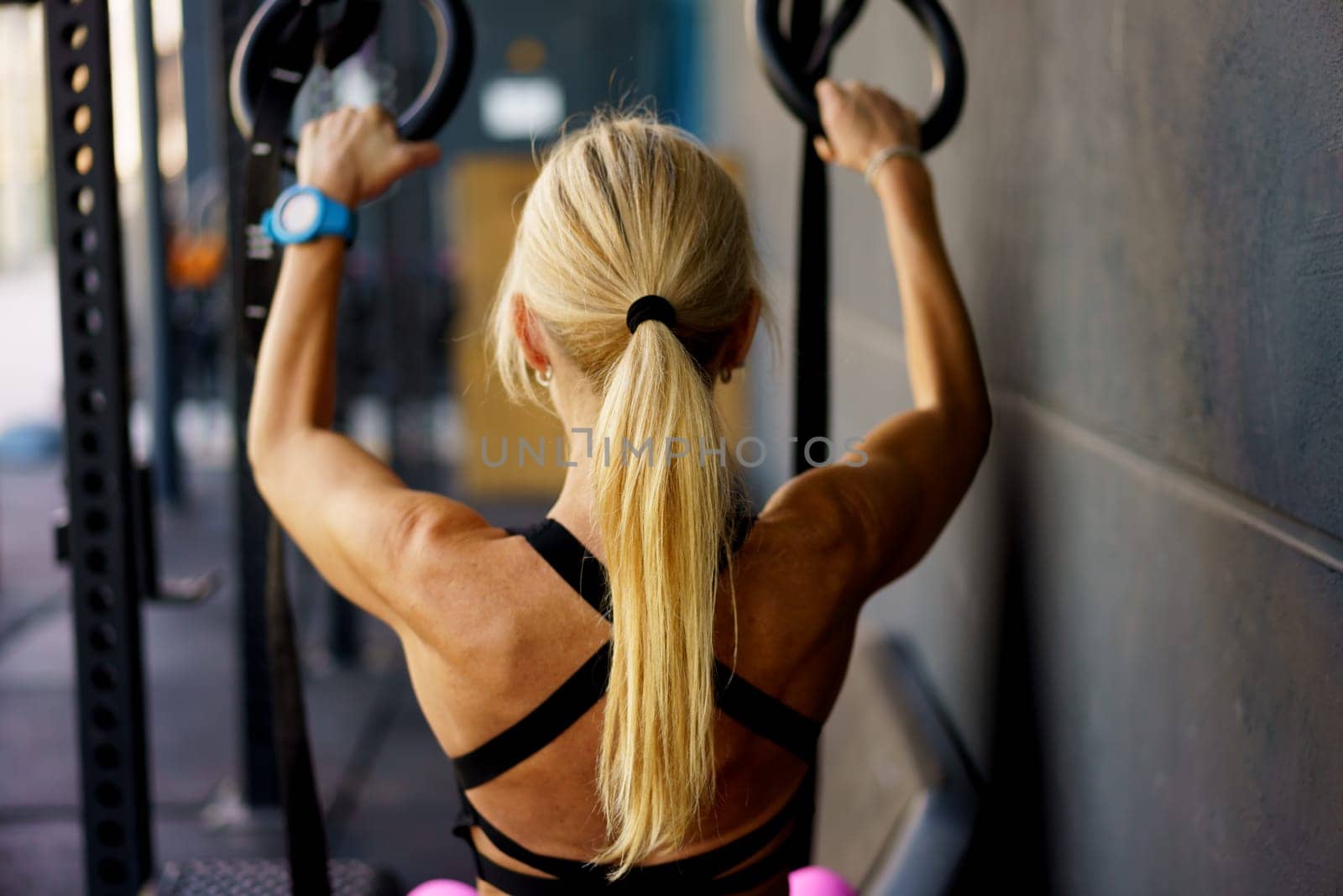 Back view of muscular female doing exercise with gymnastic rings during fitness workout in modern gym on blurred background at sport club