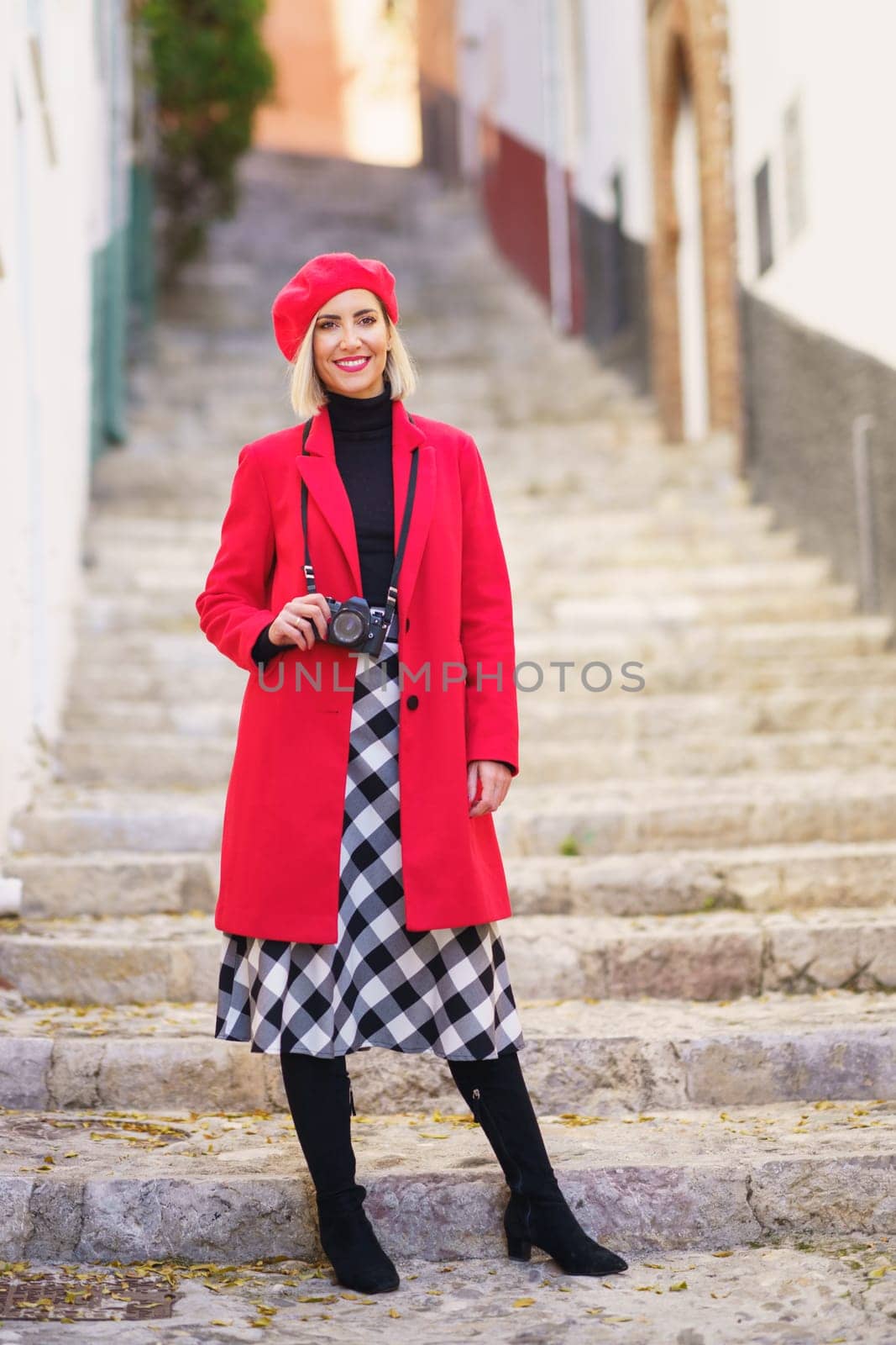 Full body of self assured young blondie, with photo camera on neck in stylish red coat and beret smiling happily while standing on stone steps between aged buildings in town