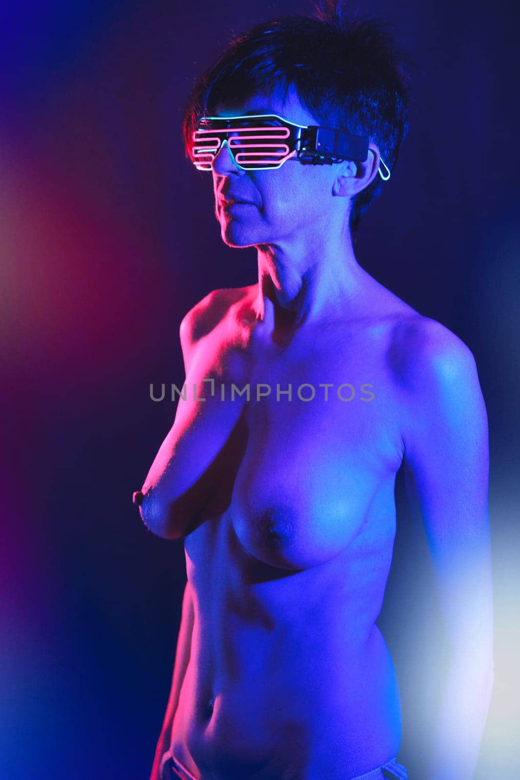 Concentrated adult female with naked breast and short dark hair in trendy neon striped shutter glasses looking away in illuminated studio