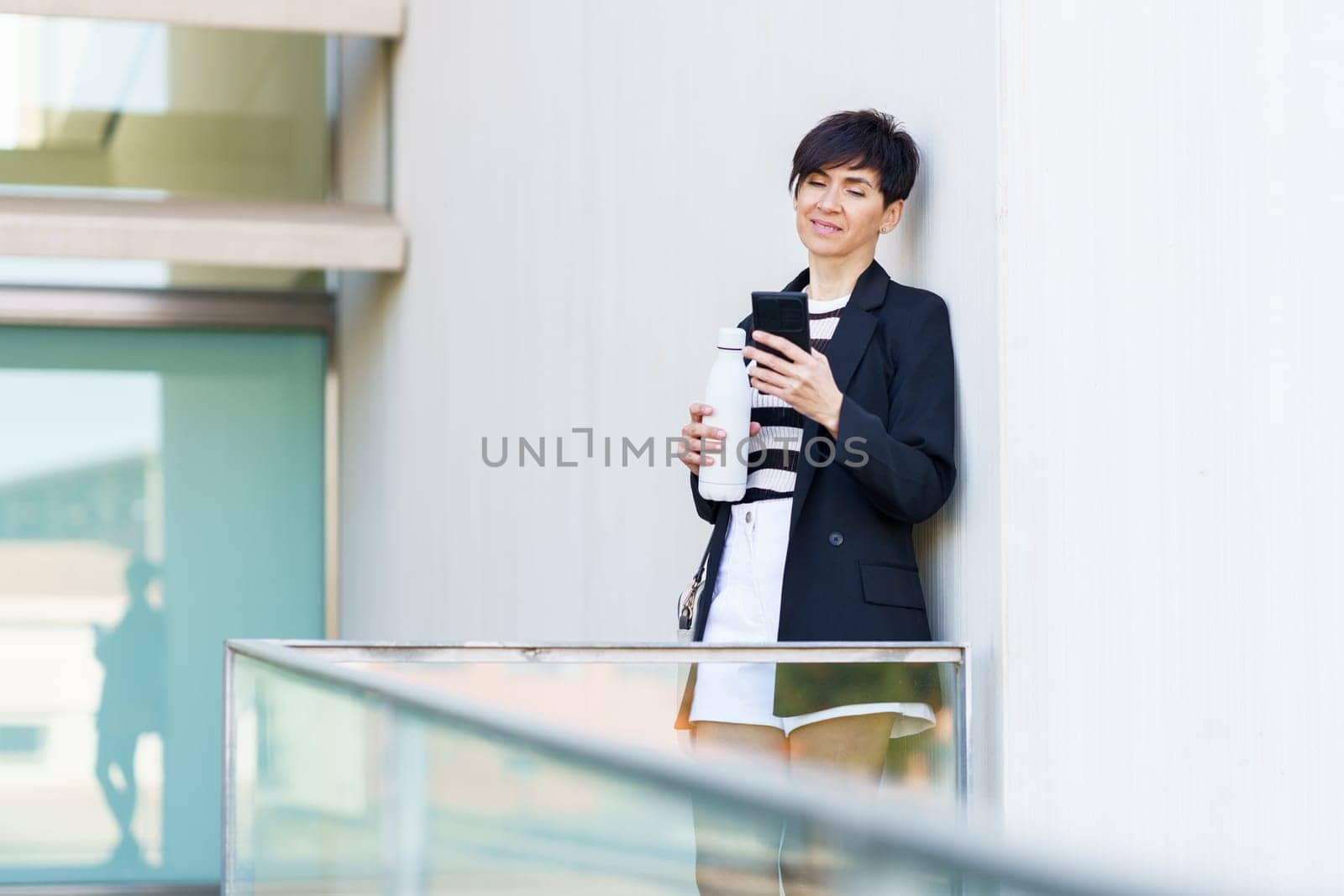 Adult businesswoman in formal clothes standing near railing and holding plastic bottle while browsing mobile phone