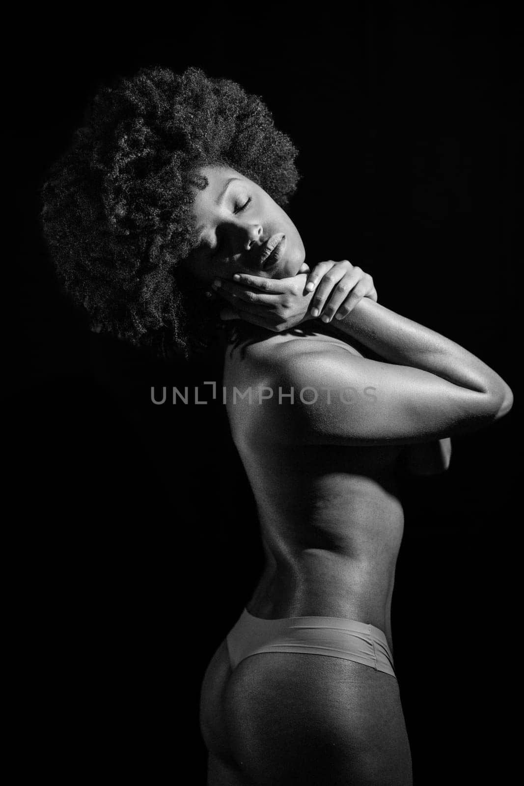 Topless black woman with Afro hairstyle closing eyes under studio light against black background. Black and white photograph.