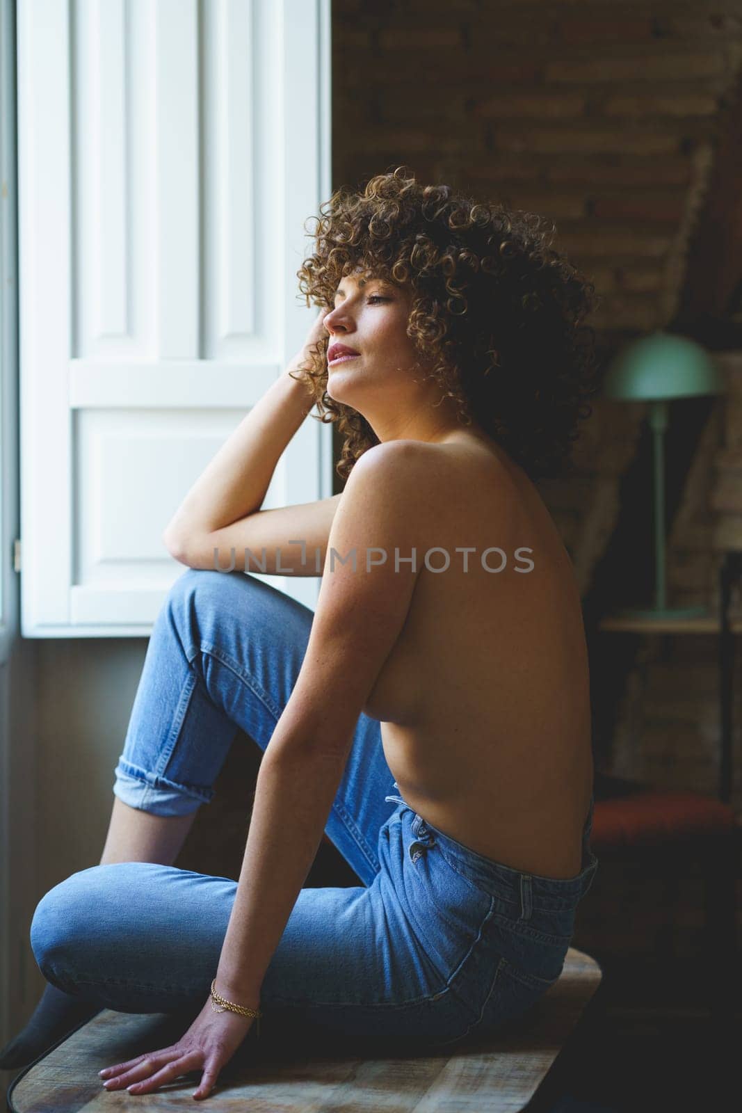 Side view of young topless female, with curly hair sitting on wooden stool with raised leg near window with shutters in room with brick wall