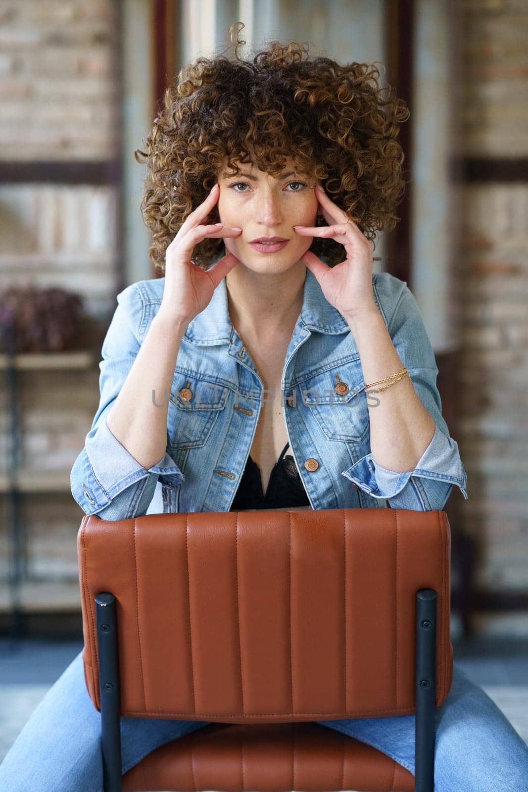 Serious young female with curly hair in denim jacket and jeans sitting on leather chair in room against brick wall while touching face