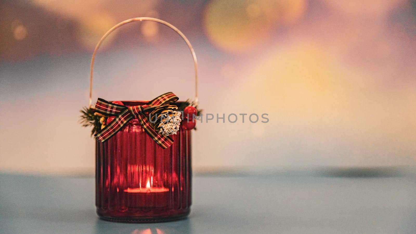 One red Christmas glass jar with a candle burning in it stands on the left on a blurred colorful background with bokke and copy space on the right, side view close-up.