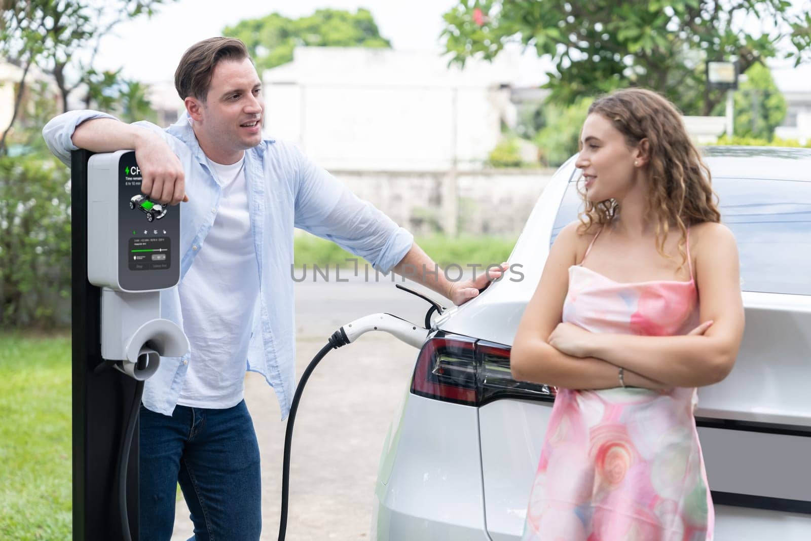 Happy and lovely couple with eco-friendly conscious recharging electric vehicle from EV charging station. EV car technology utilized as alternative transportation for future sustainability. Synchronos