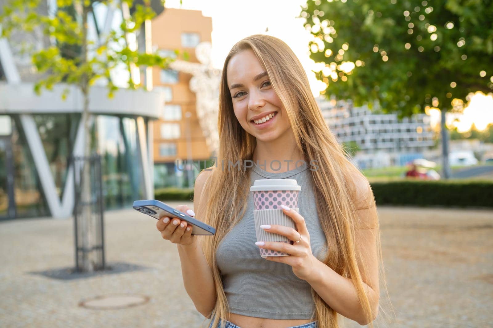 Young smiling blonde hair woman checking cell phone apps and drinks coffee standing in the street