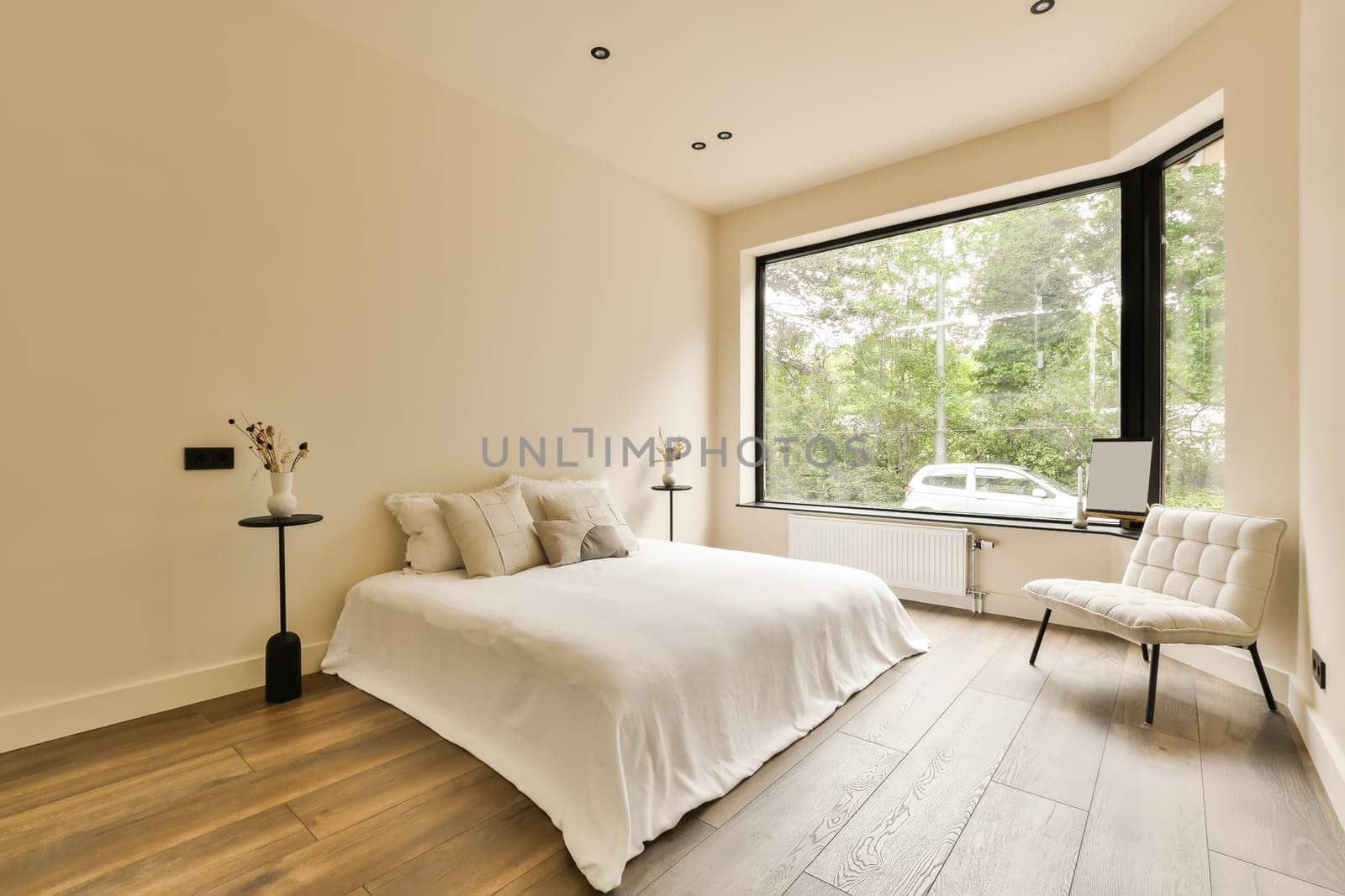 a bedroom with wood flooring and white walls, there is a large window that looks out onto the street