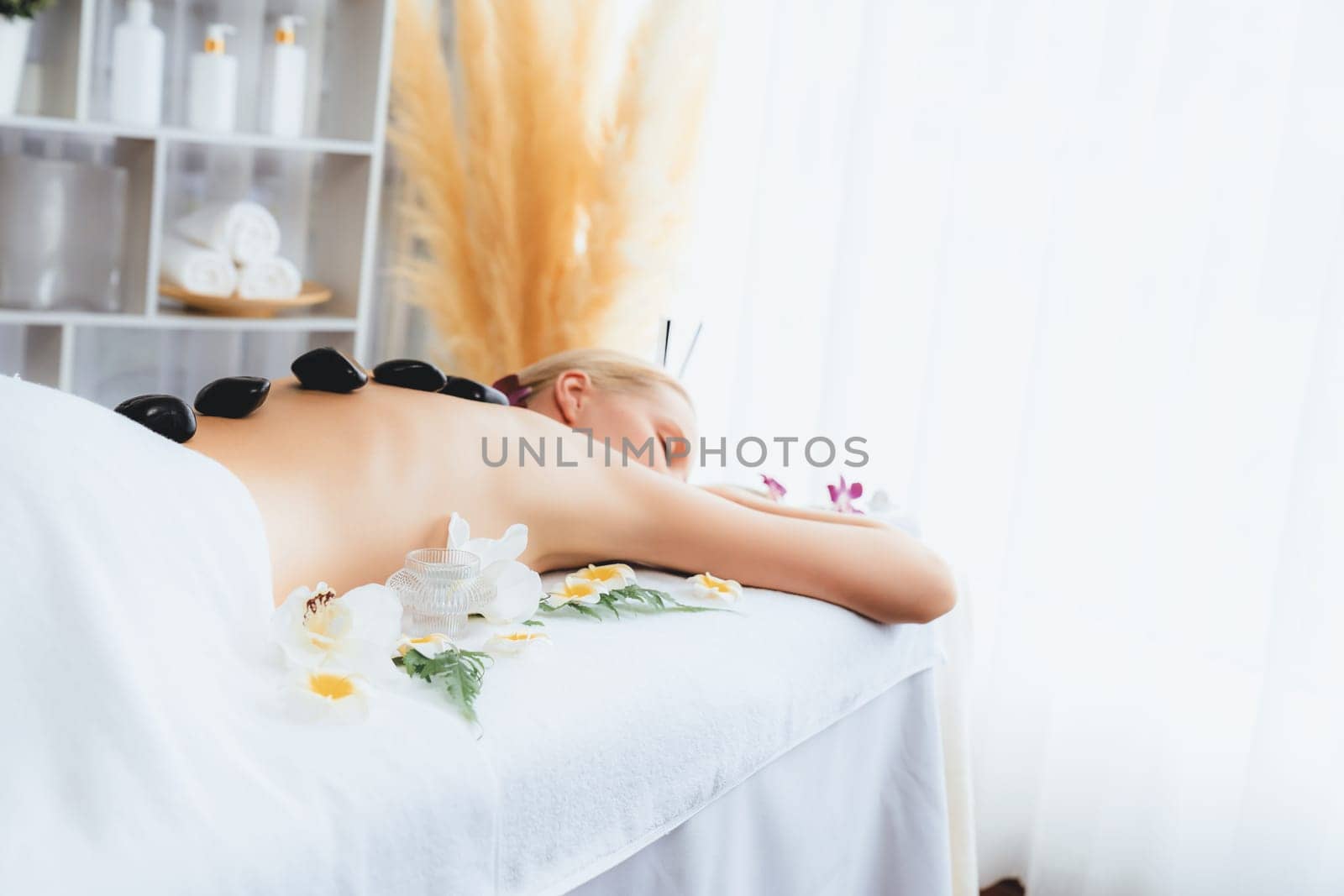 Hot stone massage at spa salon in luxury resort with day light serenity ambient, blissful woman customer enjoying spa basalt stone massage glide over body with soothing warmth. Quiescent