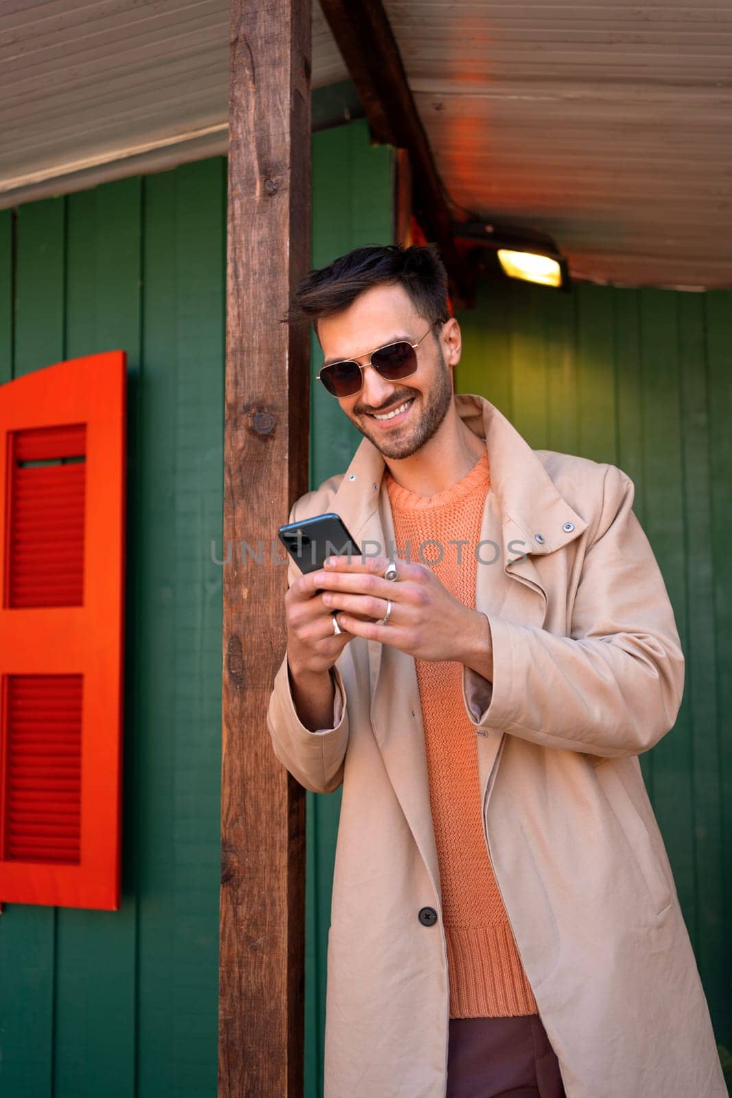 Smiling man wear sunglasses using smartphone outdoor in autumn. Stylish male standing on street leaning pole dressed stylish coat looking at his phone and laughing.
