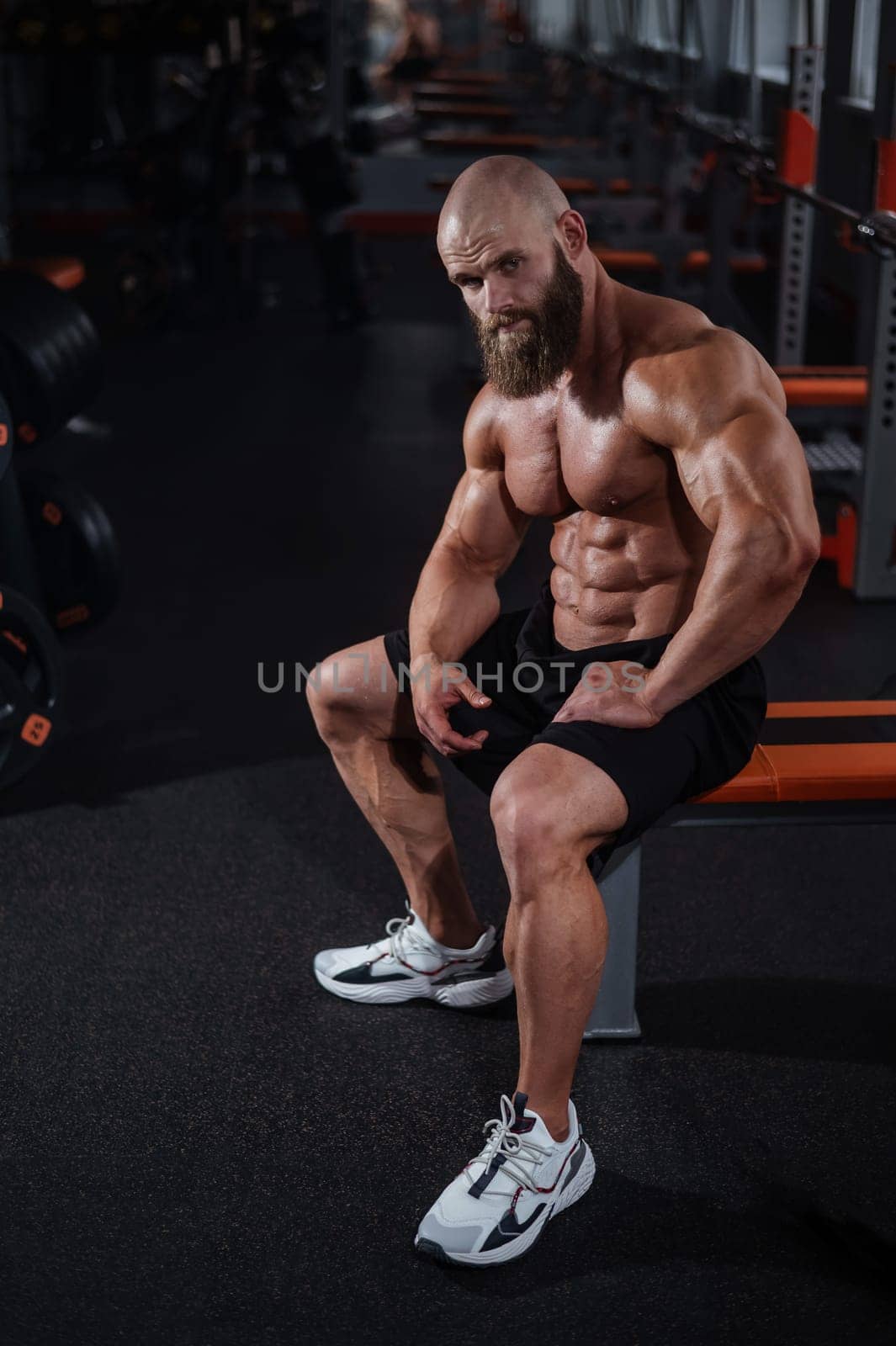 A muscular bald man in shorts is resting on a bench after a workout. Bodybuilder showing off his shape in the gym. by mrwed54