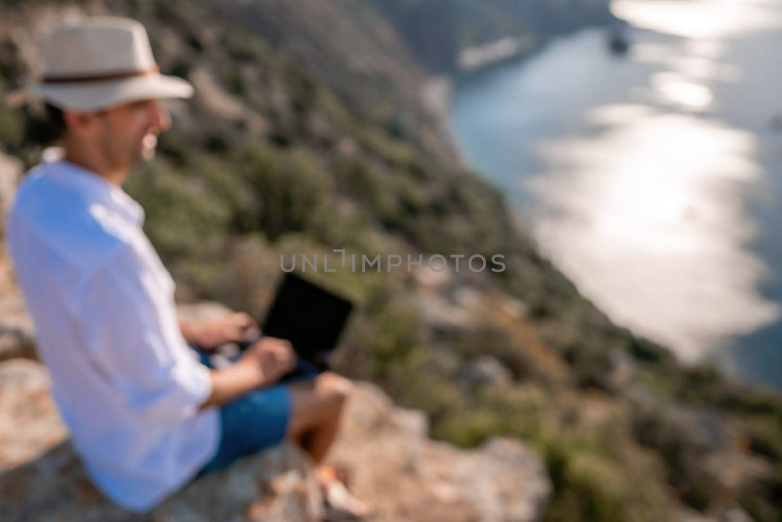 Abstract defocused/ Digital nomad, man in the hat, a businessman with a laptop sits on the rocks by the sea during sunset, makes a business transaction online from a distance. Remote work on vacation. by panophotograph