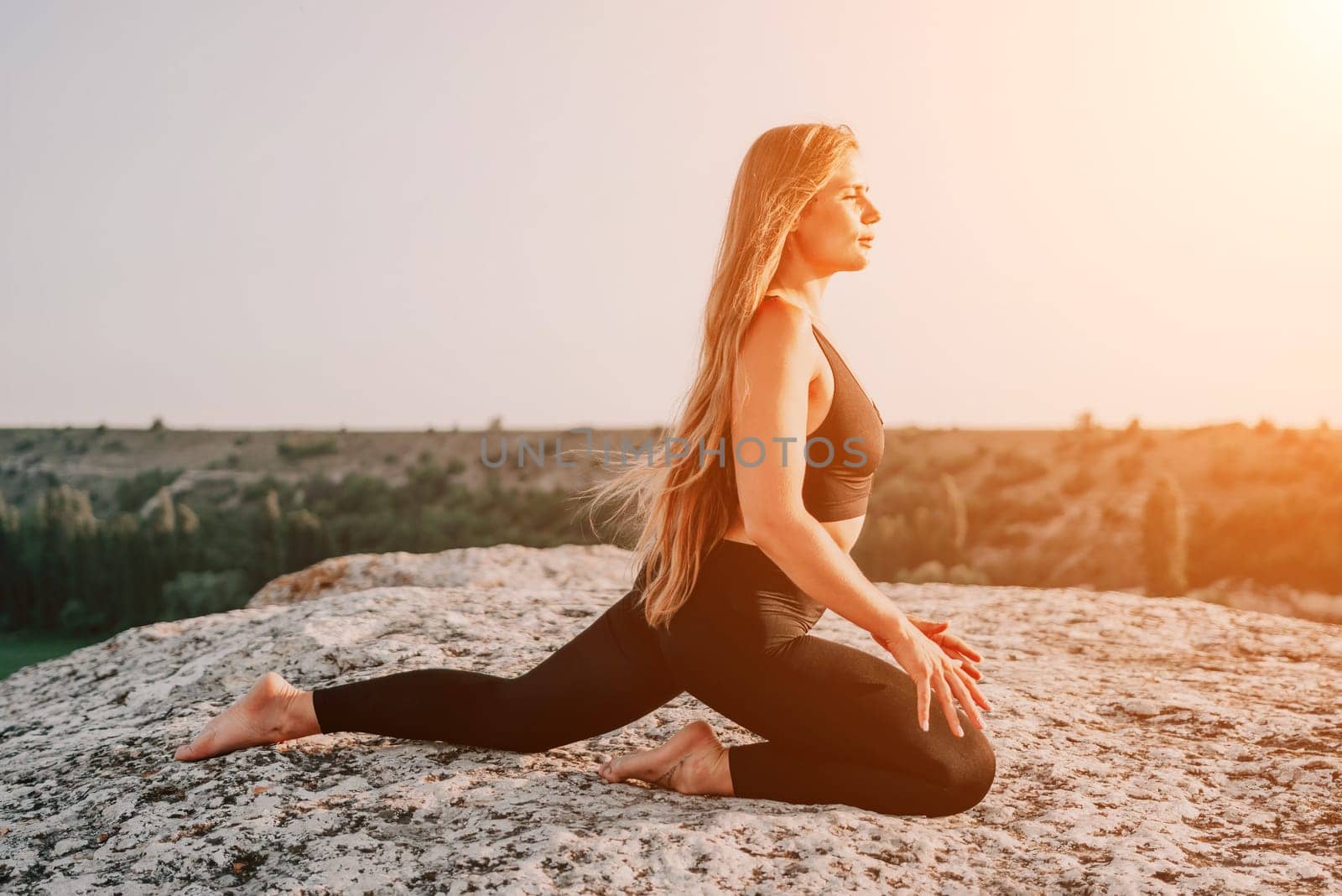 Well looking middle aged woman with long hair, fitness instructor in leggings and tops doing stretching and pilates on the rock near forest. Female fitness yoga routine concept. Healthy lifestyle.