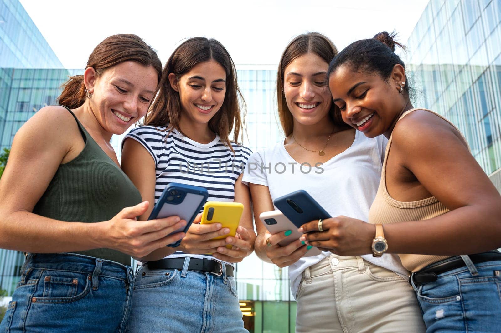 Group of happy multiracial college girl friend students looking at mobile phone ignoring each other. Smartphone and tech addiction. Youth lifestyle and social media concept.
